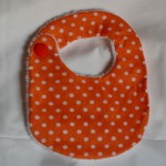 Bavoir à pois blancs sur fond orange, doublé de tissu éponge blanc, 100% coton, fermé par un gros bouton. Lavage 30Â° en machine.